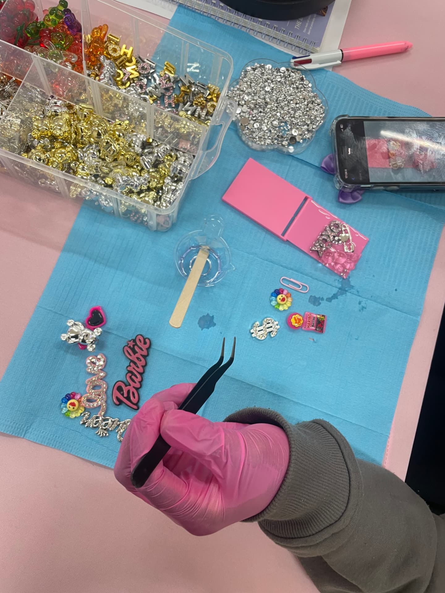 Overhead view of a craft desk: a hand in a pink glove uses tweezers to place small charms from a tray of gems, letters and beads onto a decorated phone case