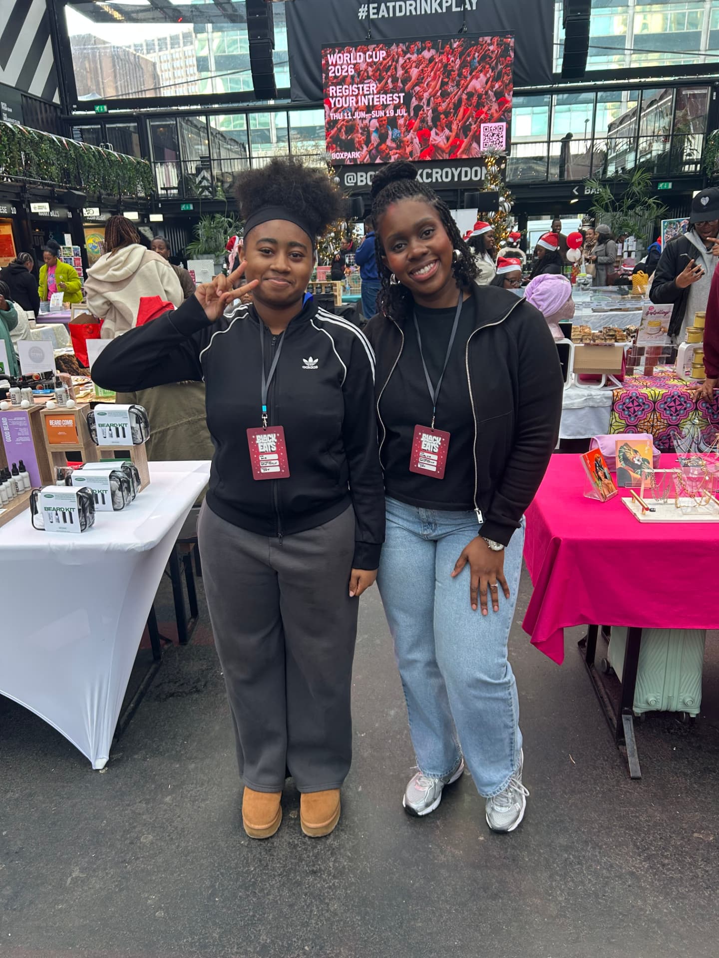 Two young people wearing Black Eats LDN event badges stand together between market stalls at BOXPARK Croydon