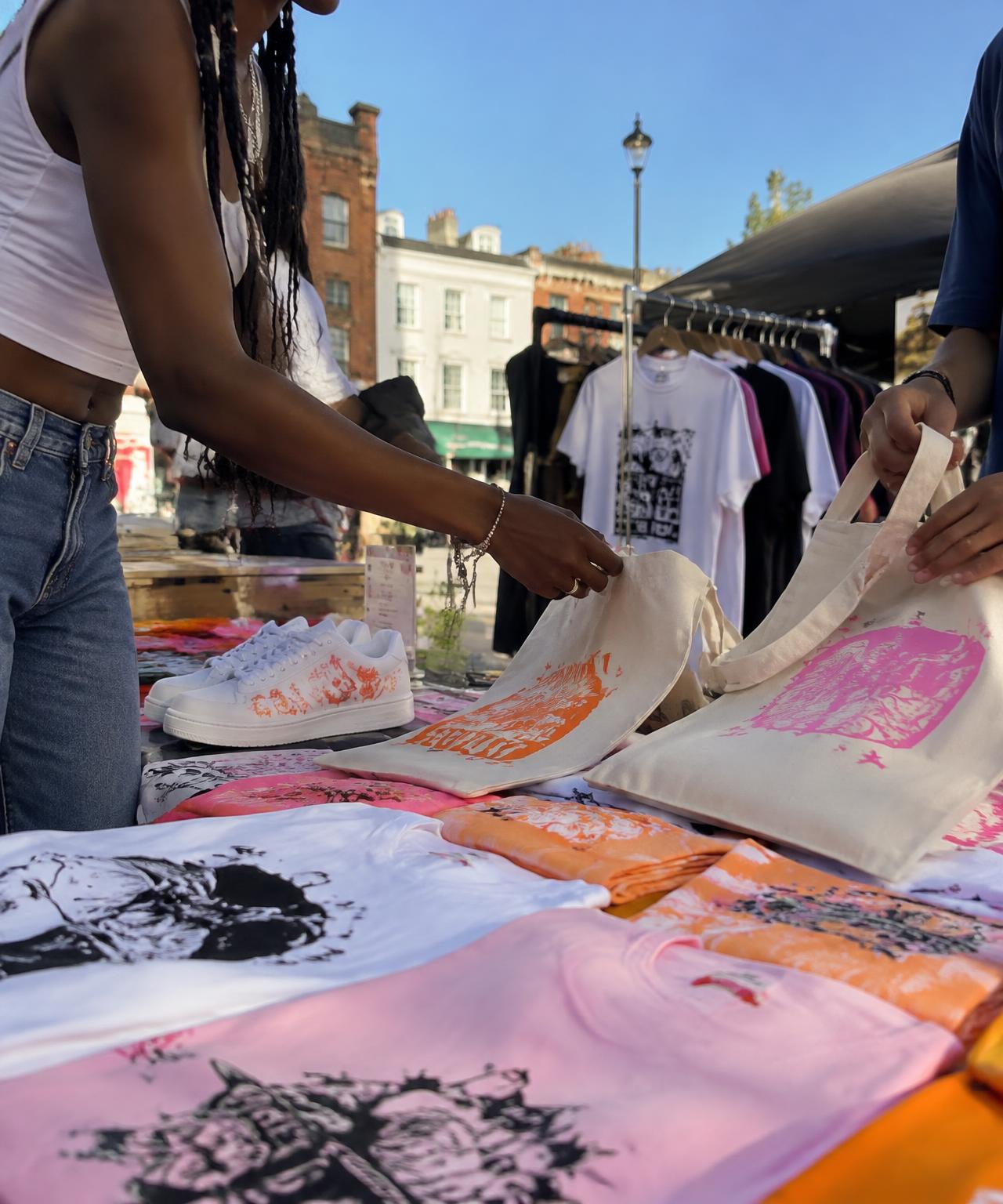 Young people arranging hand-printed tote bags and t-shirts at a London pop-up market stall